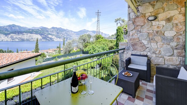 Ein Balkon mit Steinmauer, Korbmöbeln, einem Tisch mit Flasche und Gläsern sowie Blick auf Berge und See.