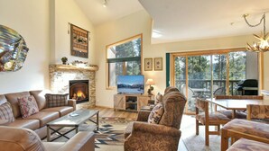 A spacious living room with a fireplace, comfortable seating, a television, and a large window offering a view of the outdoors. - Chimney Ridge Larkspur Townhome: Hot Tub, Great Location (Breckenridge)