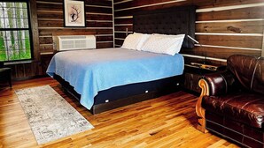 A bedroom with a bed, a brown leather sofa, wooden panel walls, and a window with a view of trees. - {Big Timbers} Lodging next to North Entrance of Itasca State Park (Shevlin)