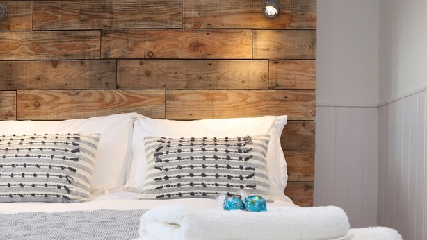 Una cama bien tendida con sábanas blancas y almohadas a rayas, un cabecero de madera y una lámpara de noche pequeña.
