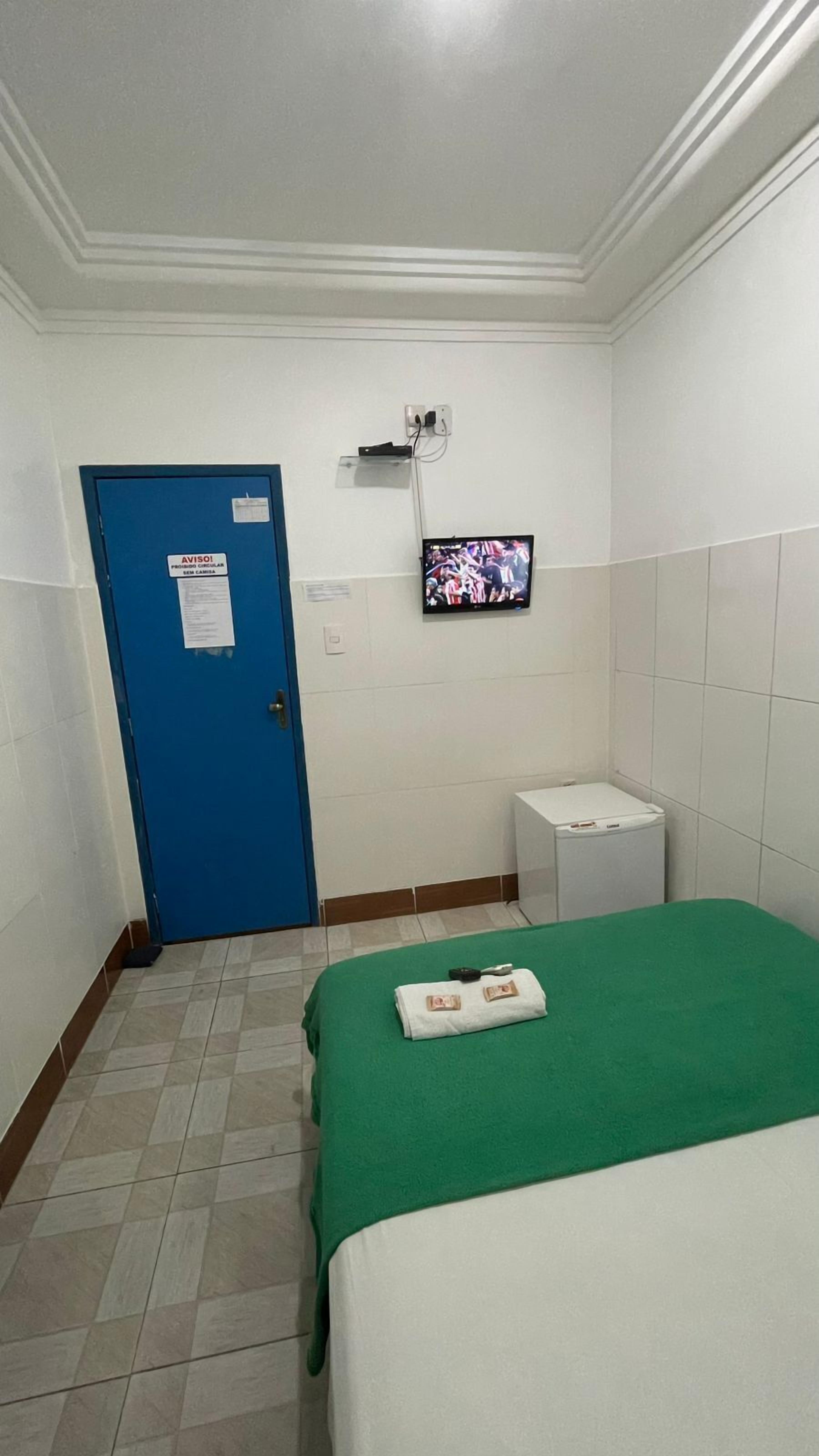 A hotel room with a single bed, a small bedside table, a TV mounted on the wall, and a blue door with a notice.