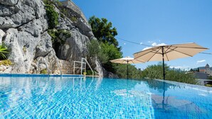 Piscina azul cristalina com um fundo rochoso, uma escada e guarda-sóis.