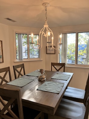 A dining area with a wooden table, chairs, a chandelier, and windows with views of trees and buildings.