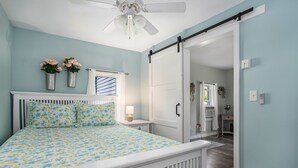 Une chambre à coucher comprenant un lit, une table de chevet, un vase fixé au mur avec des fleurs, un ventilateur de plafond et une porte coulissante donnant sur une autre pièce.