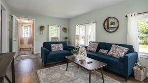 Un salon avec un canapé bleu foncé, une table basse et un tapis. Il y a deux fenêtres avec des rideaux, un miroir fixé au mur et une porte qui donne sur une autre pièce.