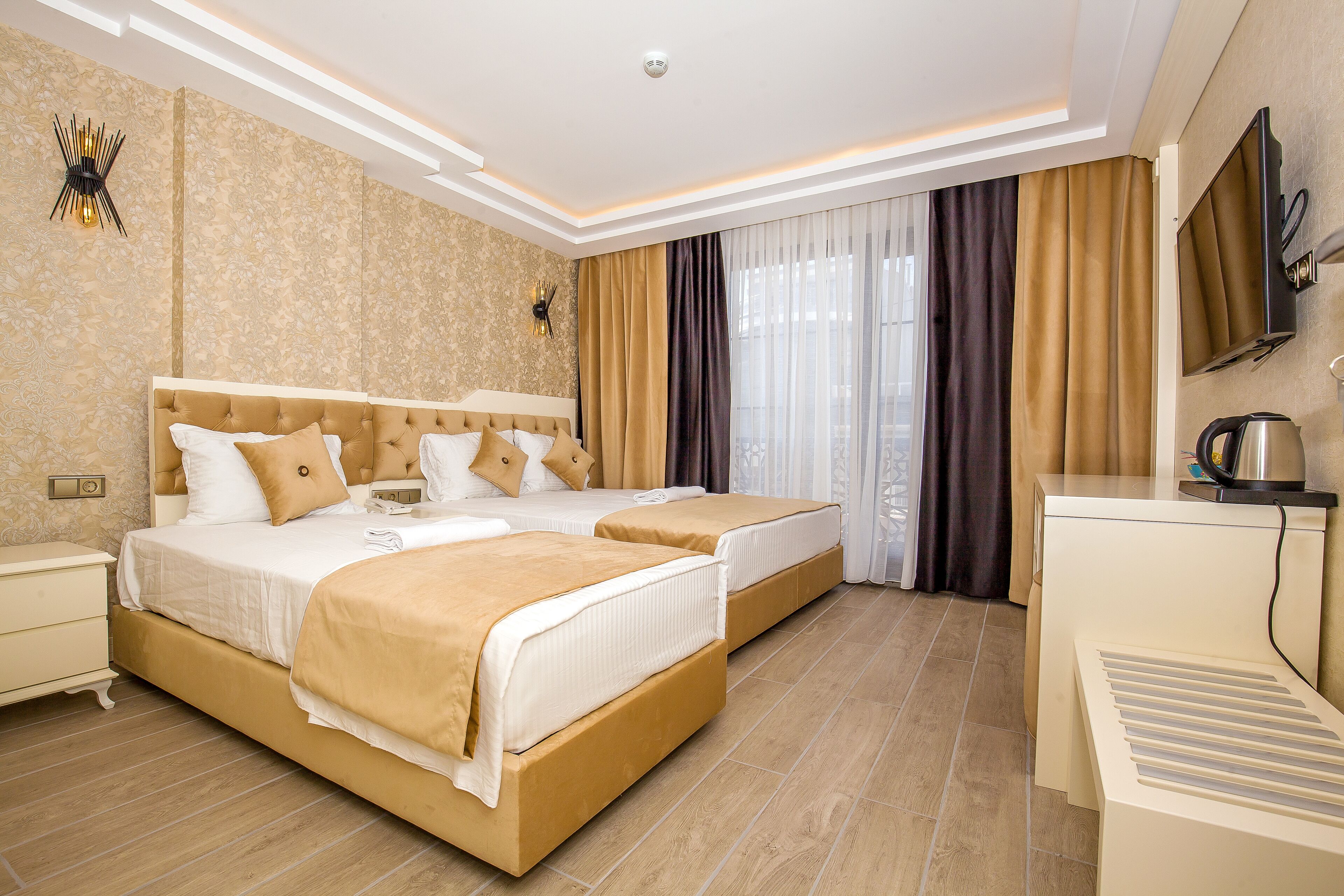 Habitación de hotel con dos camas, televisión, escritorio con hervidor y un amplio ventanal con cortinas.