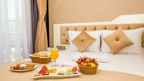 Una cama bien hecha con cabecera, una cesta de frutas, una bandeja con alimentos para el desayuno y un vaso de jugo de naranja sobre una mesa de habitación de hotel.