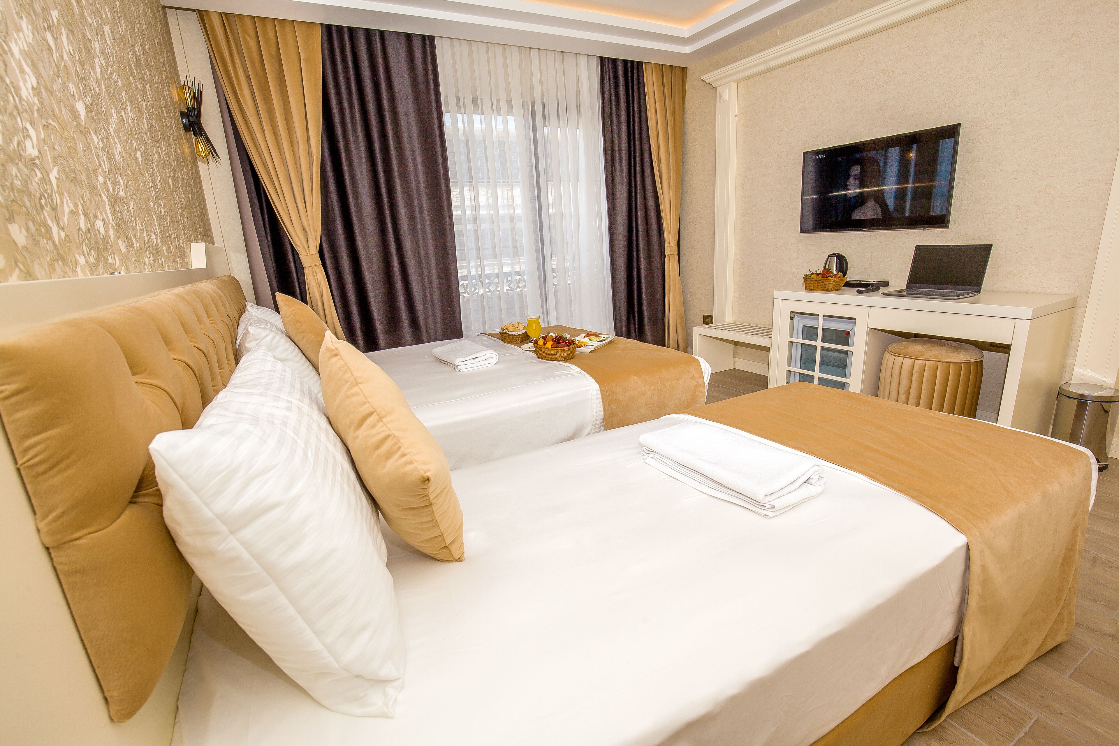 A hotel room with two beds, a TV, a desk with a laptop, and a window with curtains.
