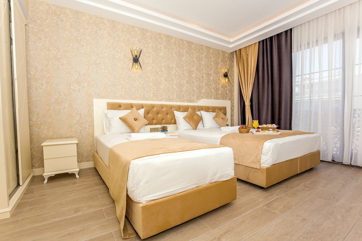 Quarto de hotel com duas camas, uma mesinha de cabeceira, uma luminária na parede e uma janela com cortinas.