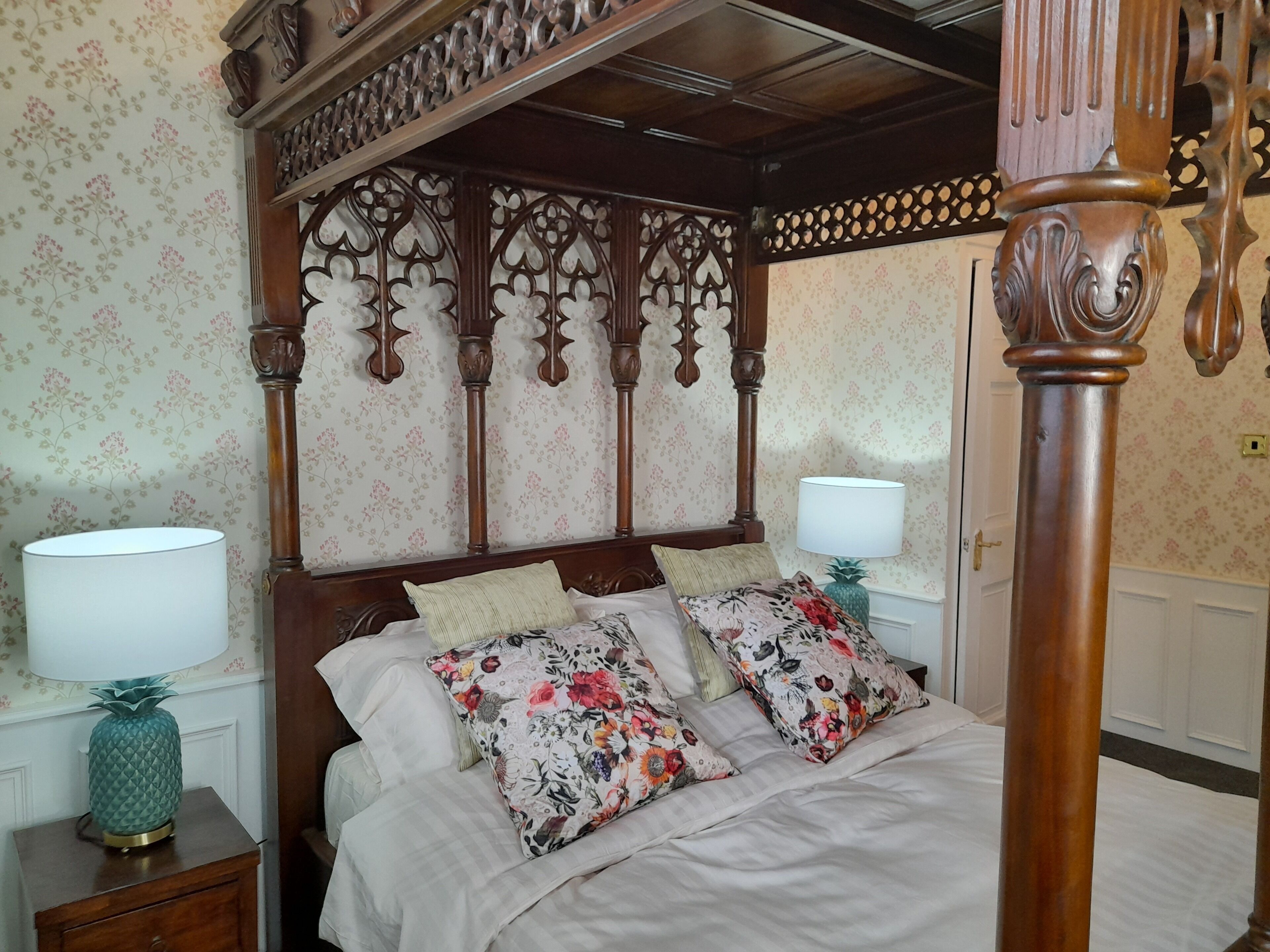 A four-poster bed with floral-patterned pillows, a wooden headboard, and a pineapple-shaped nightstand lamp.