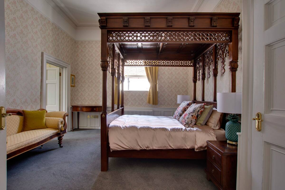 Ein Zimmer mit einem Himmelsbett, einer Sitzecke, einem Nachttisch und einer Heizung, dessen Wände mit einem Muster-Tapete versehen sind.