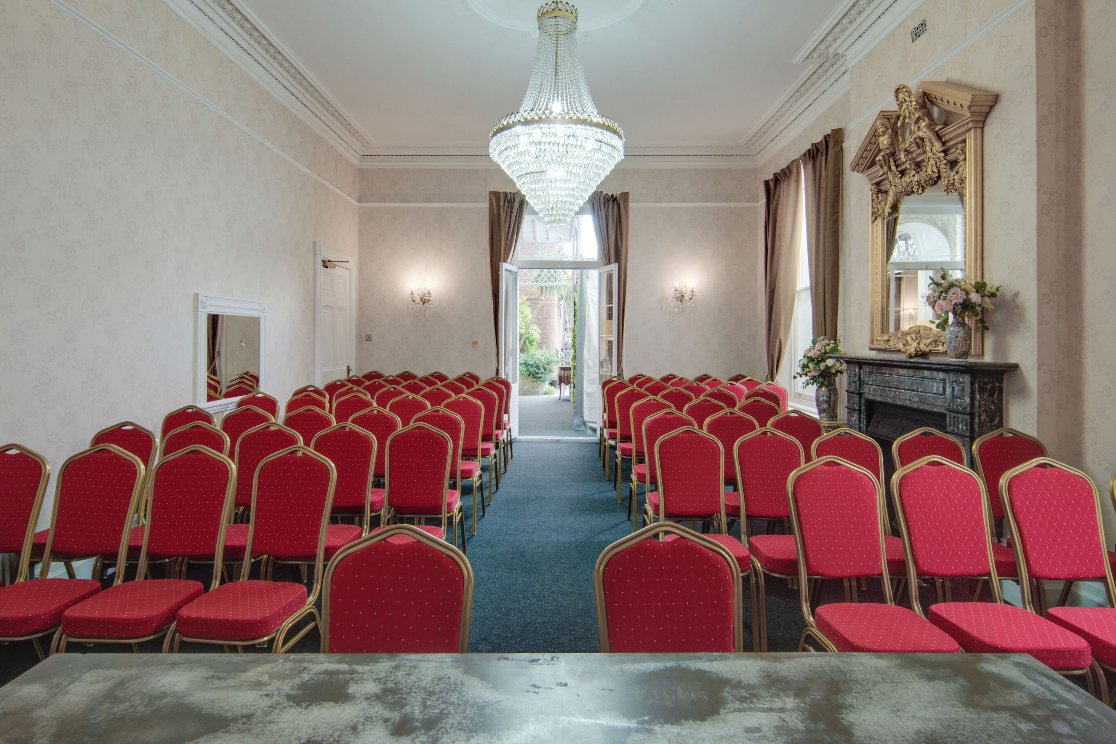 A conference room with rows of red chairs, a chandelier, and a fireplace with a mirror and vase.