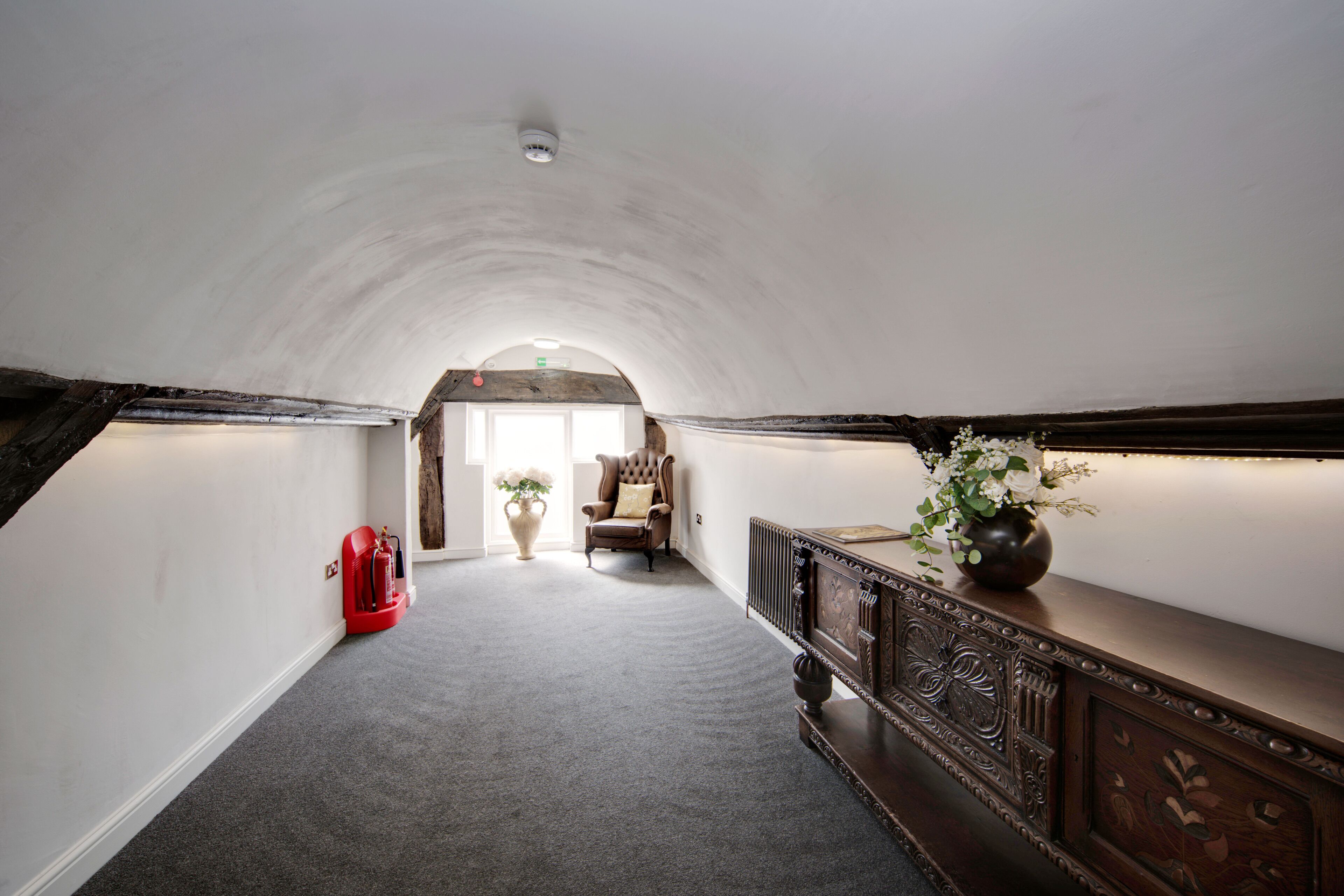 A hallway with a vaulted ceiling, a fireplace, a red fire extinguisher, and a vase with flowers.