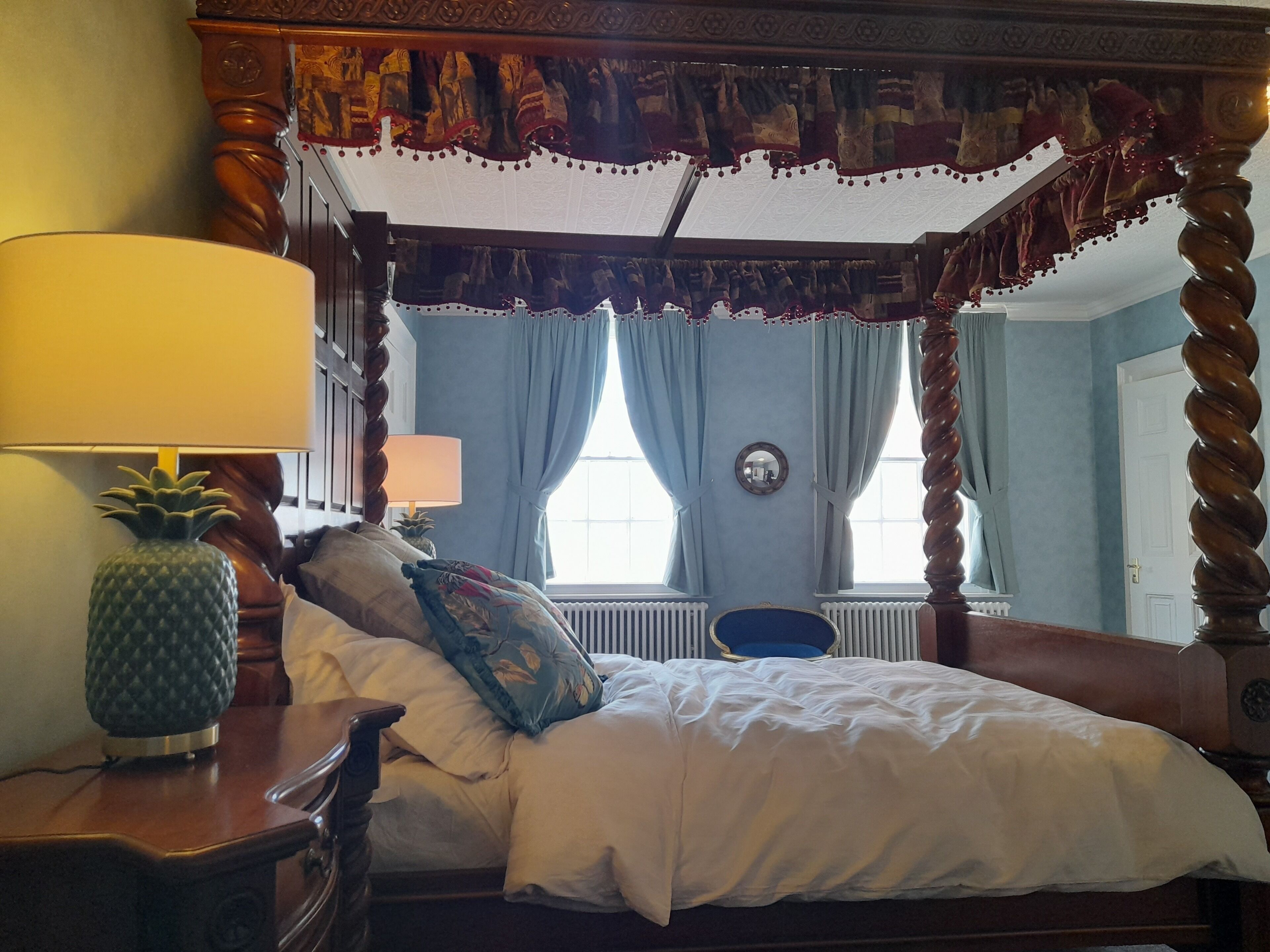A four-poster canopy bed with a mirror above it, a pineapple-shaped lamp, and a blue chair in a room with light blue walls and a white door.