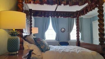 A four-poster canopy bed with a mirror above it, a pineapple-shaped lamp, and a blue chair in a room with light blue walls and a white door.