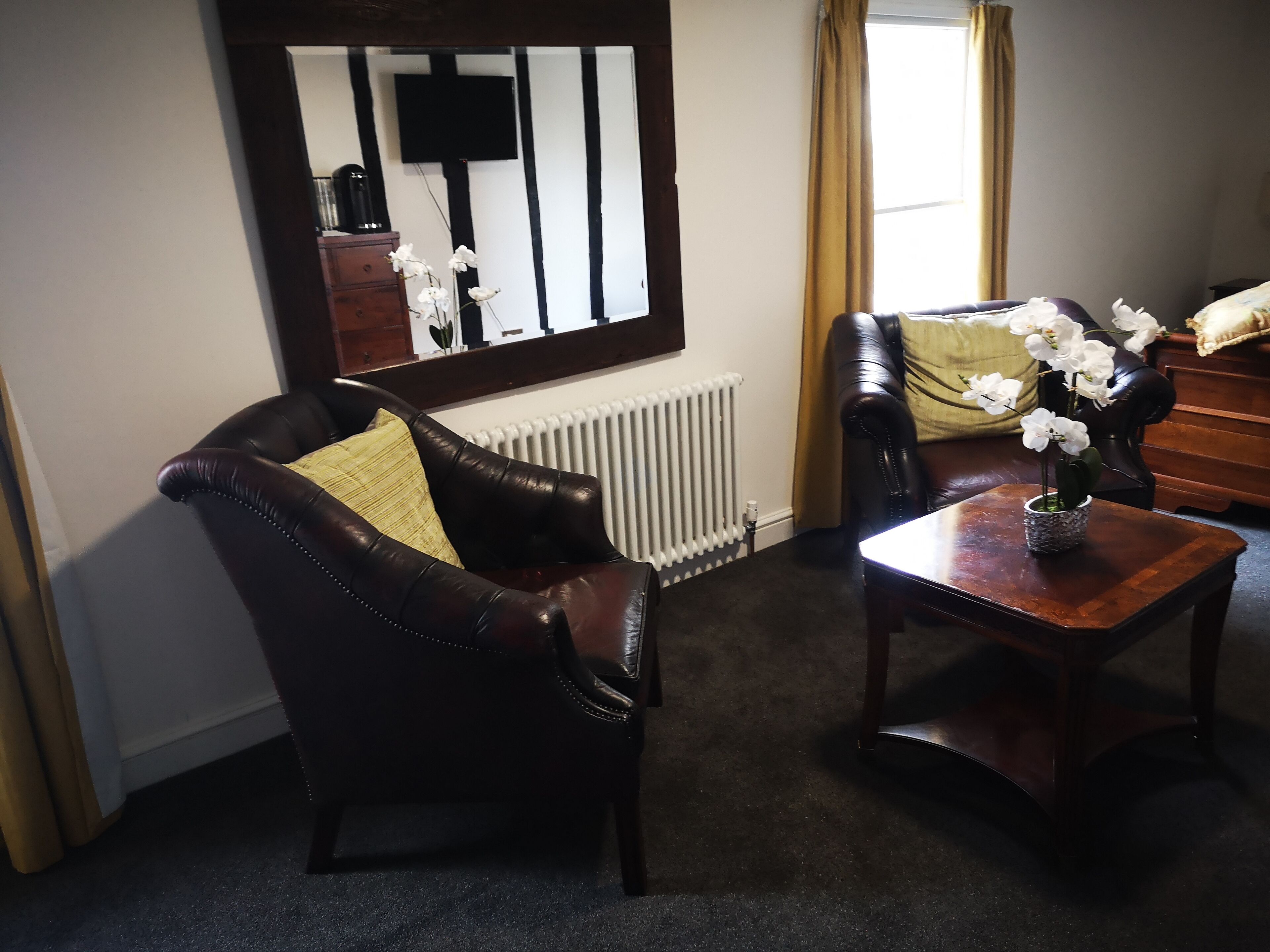 Une pièce comprenant un fauteuil en cuir marron foncé, un canapé assorti, une petite table en bois sur laquelle est posé un vase de fleurs blanches, un radiateur et un grand miroir accroché au mur.