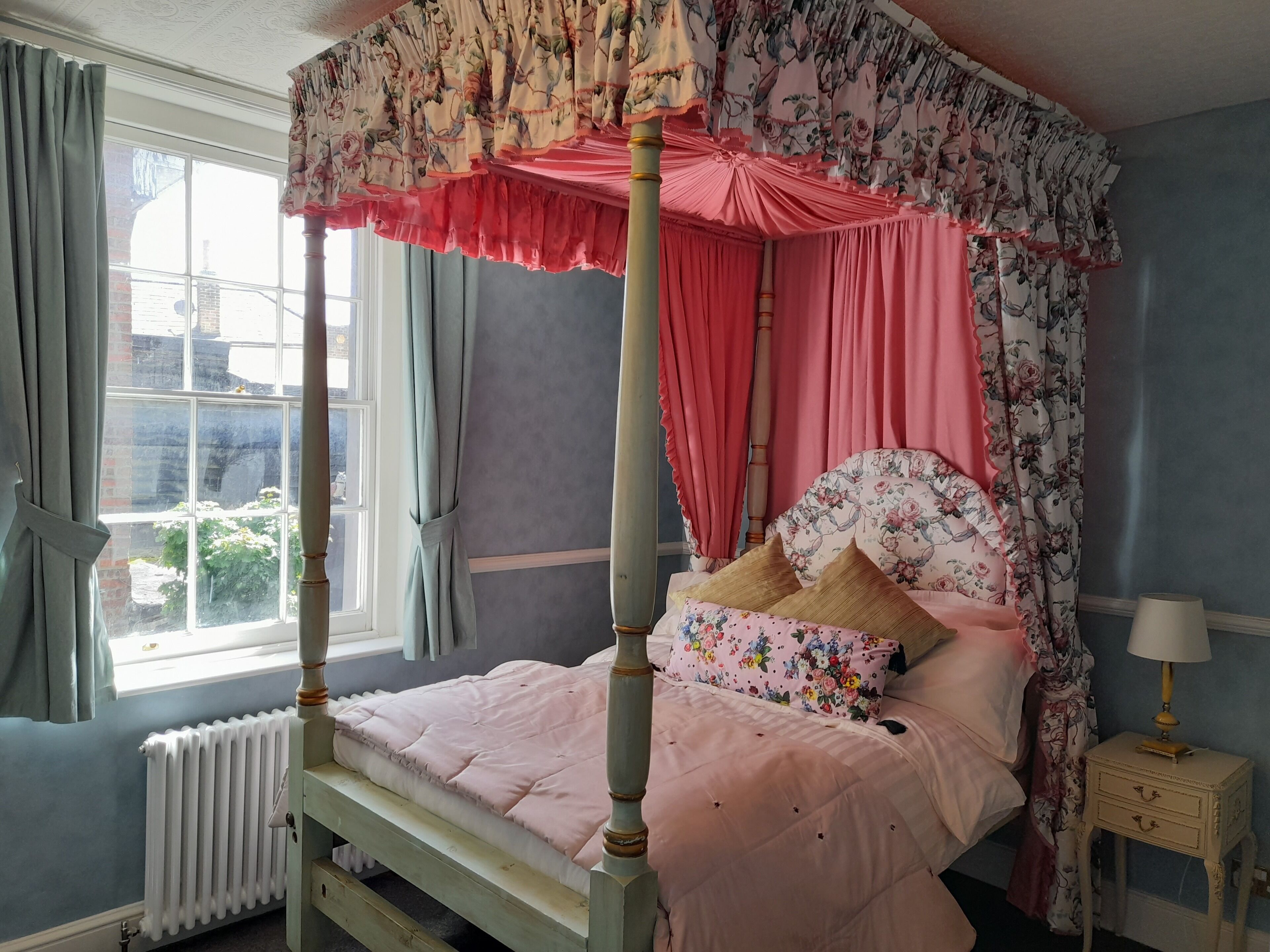 A four-poster bed with floral curtains, a headboard with floral patterns, and a bedside table with a lamp.