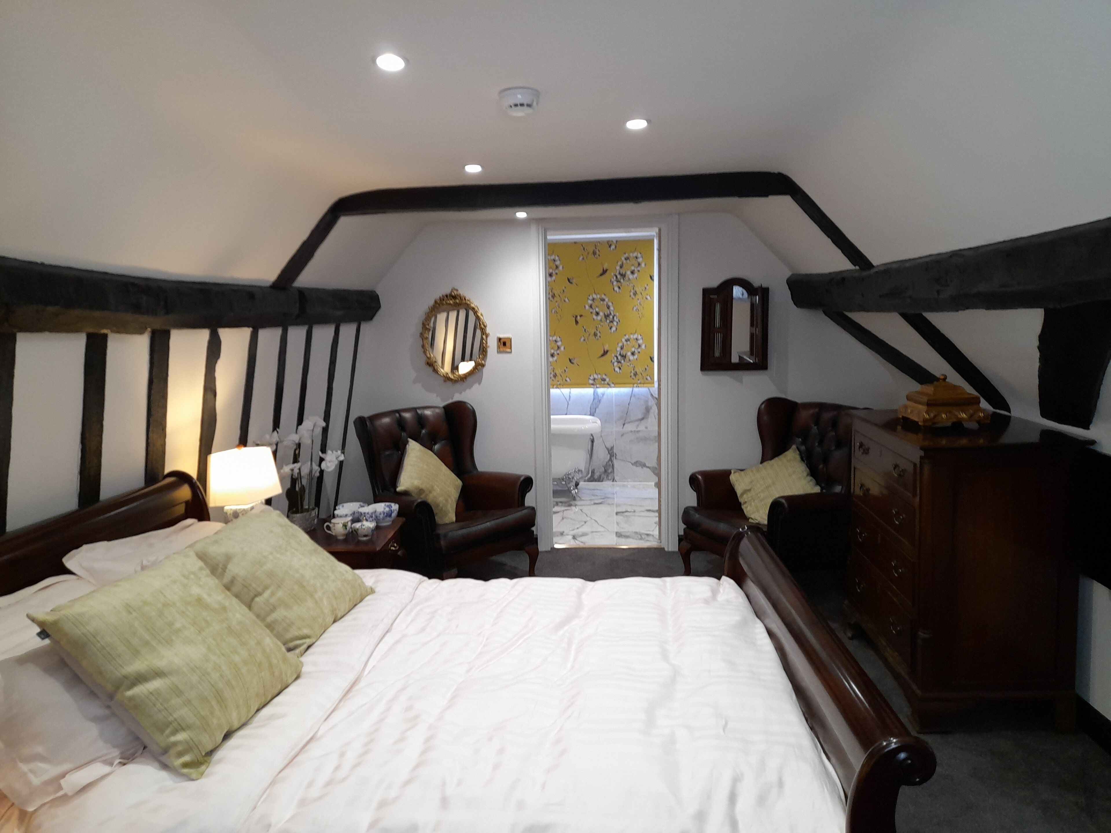 A bedroom with a large bed, two armchairs, a dresser, and a mirror on the wall.