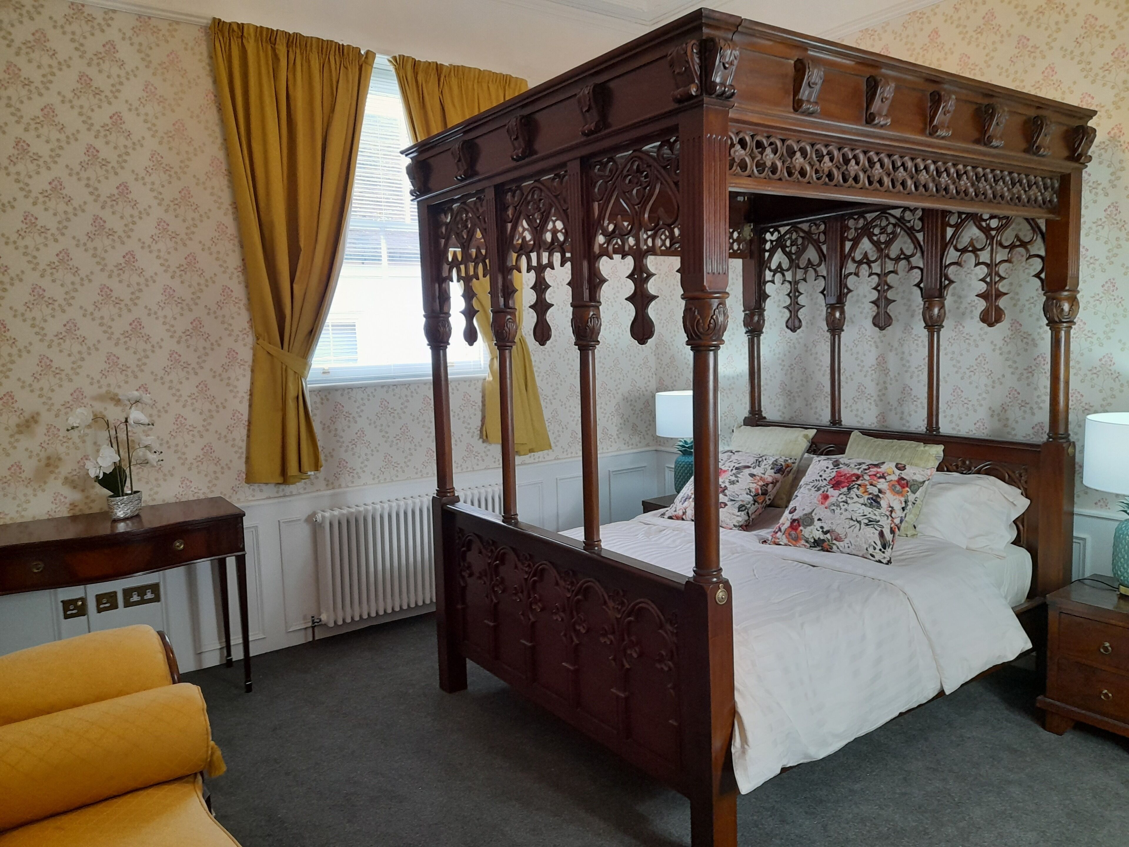 A four-poster bed with intricate carvings, a floral-patterned wall, a radiator, and a yellow armchair.