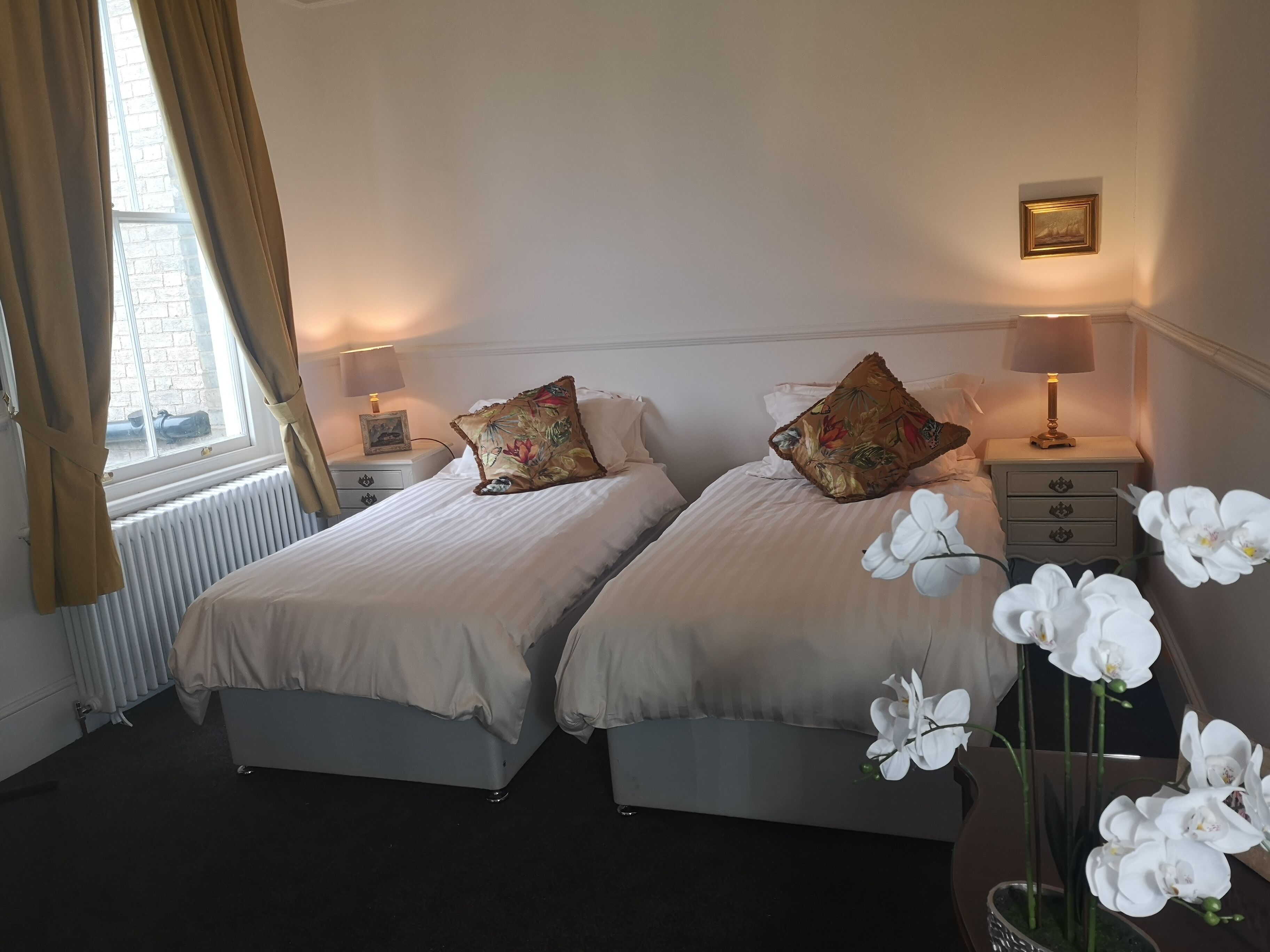 A hotel room with two beds, a window with curtains, bedside lamps, a framed picture, and a vase with white flowers.