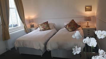 A hotel room with two beds, a window with curtains, bedside lamps, a framed picture, and a vase with white flowers.