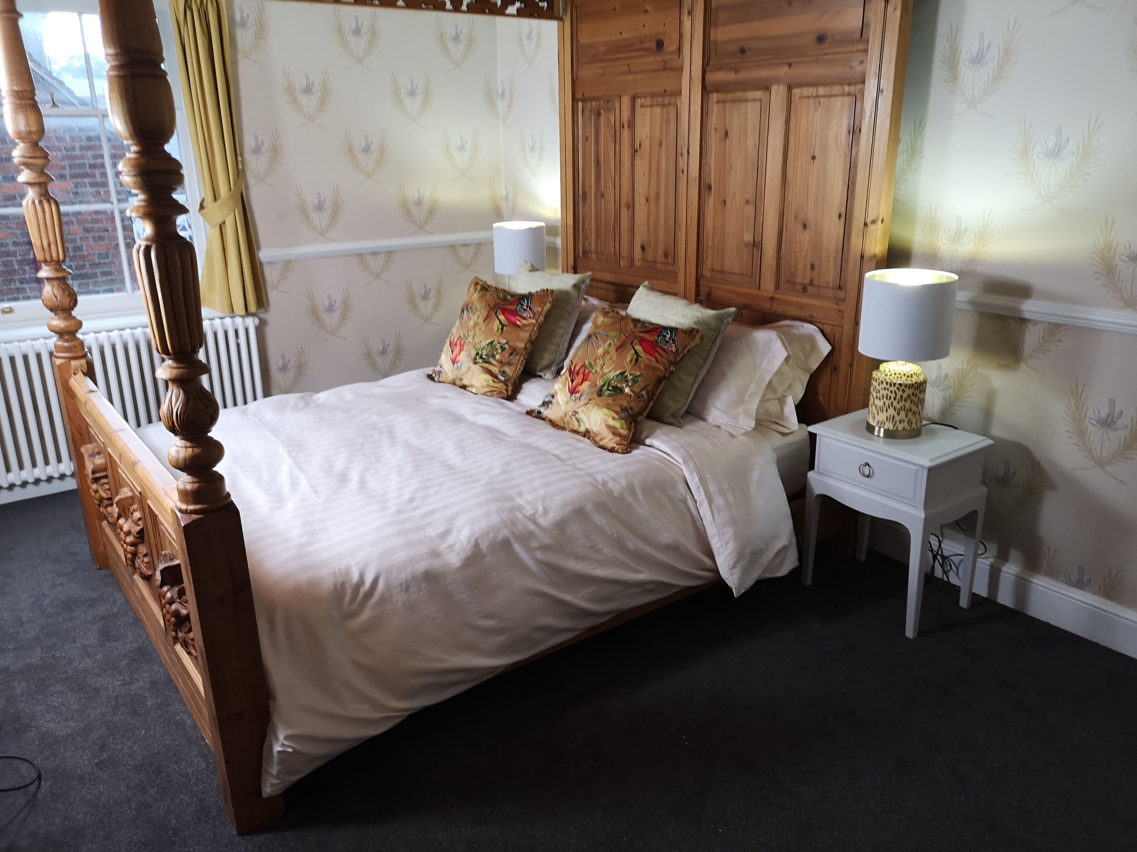 Ein Schlafzimmer mit einem hölzernen Himmelbett, zwei Nachttischlampen und blumenmusterter Tapete.
