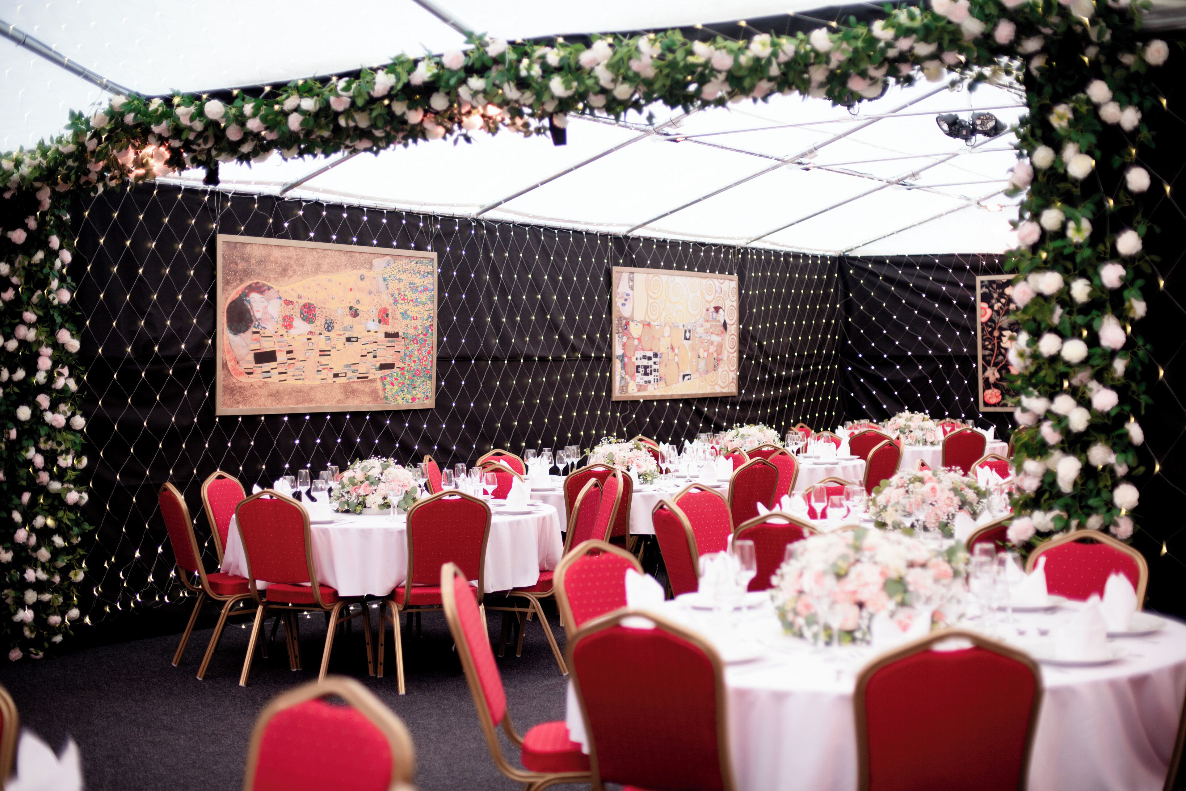 A tented event space with round tables, red chairs, and floral decorations.