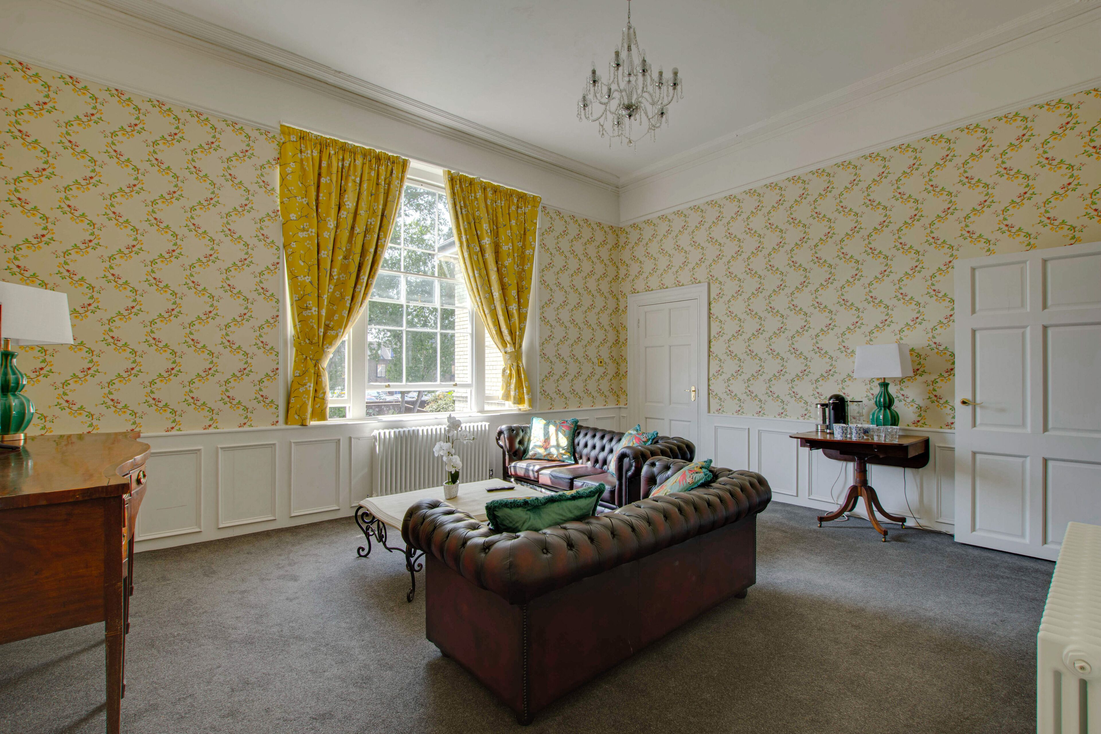 A room with a brown leather sofa, a wooden table, a radiator, and a window with yellow curtains.