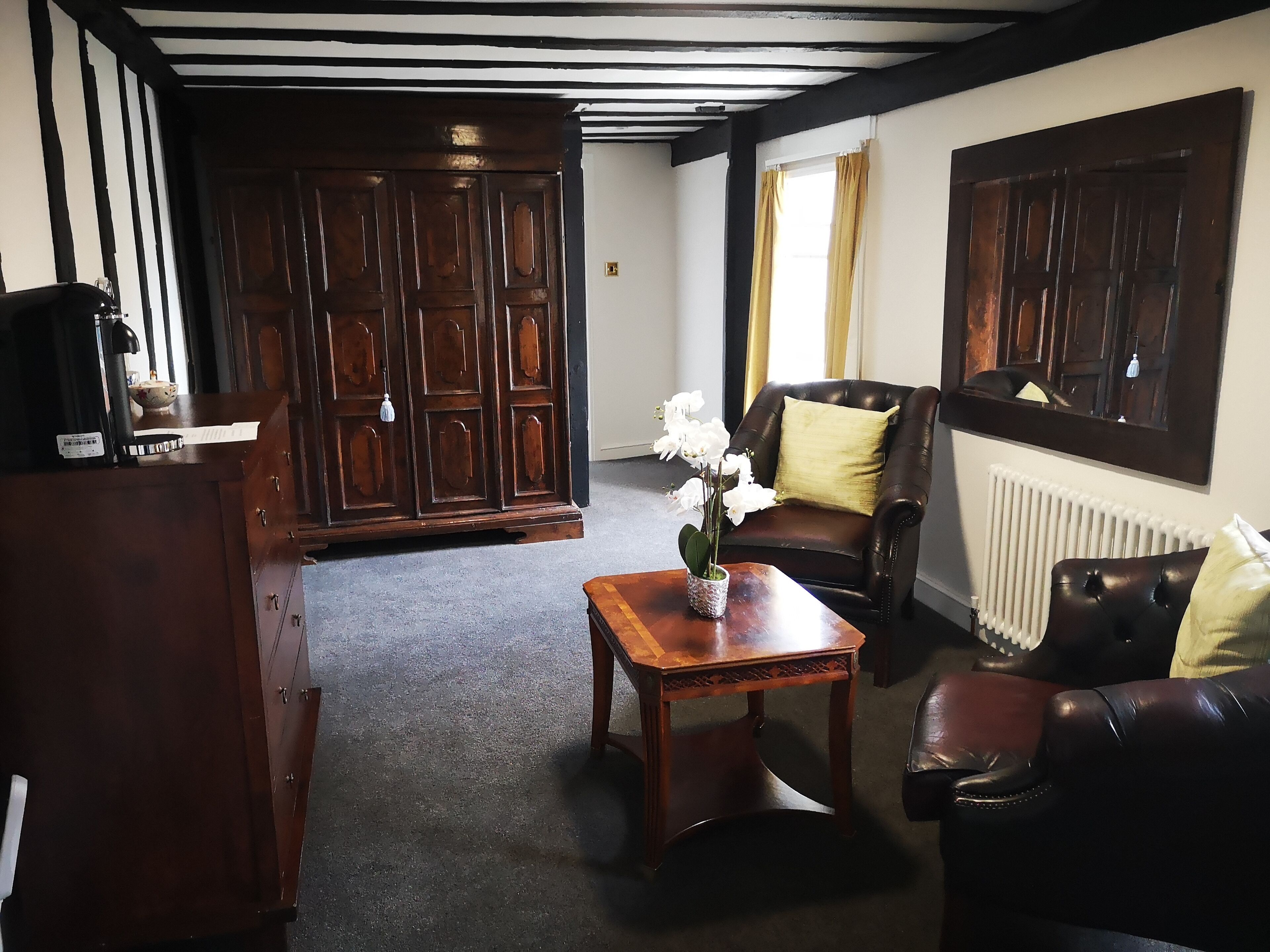 Ein Zimmer mit einem dunklen Holzschrank, einem Ledersessel mit goldenem Kissen, einem Holztisch mit einer Vase voller weißer Blumen und einem Heizkörper an der Wand.