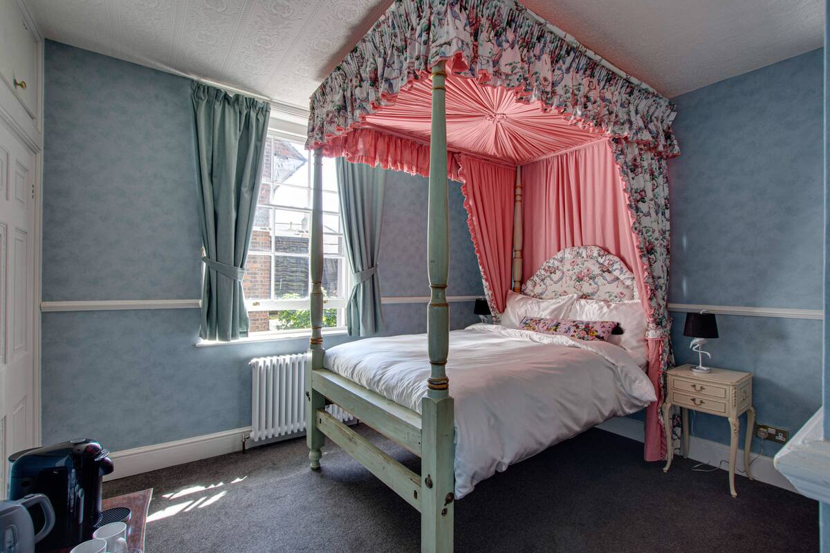 Ein Himmelsbett mit Vorhängen mit Blumenmuster, ein Nachttisch mit Lampe und ein Fenster mit Vorhängen.