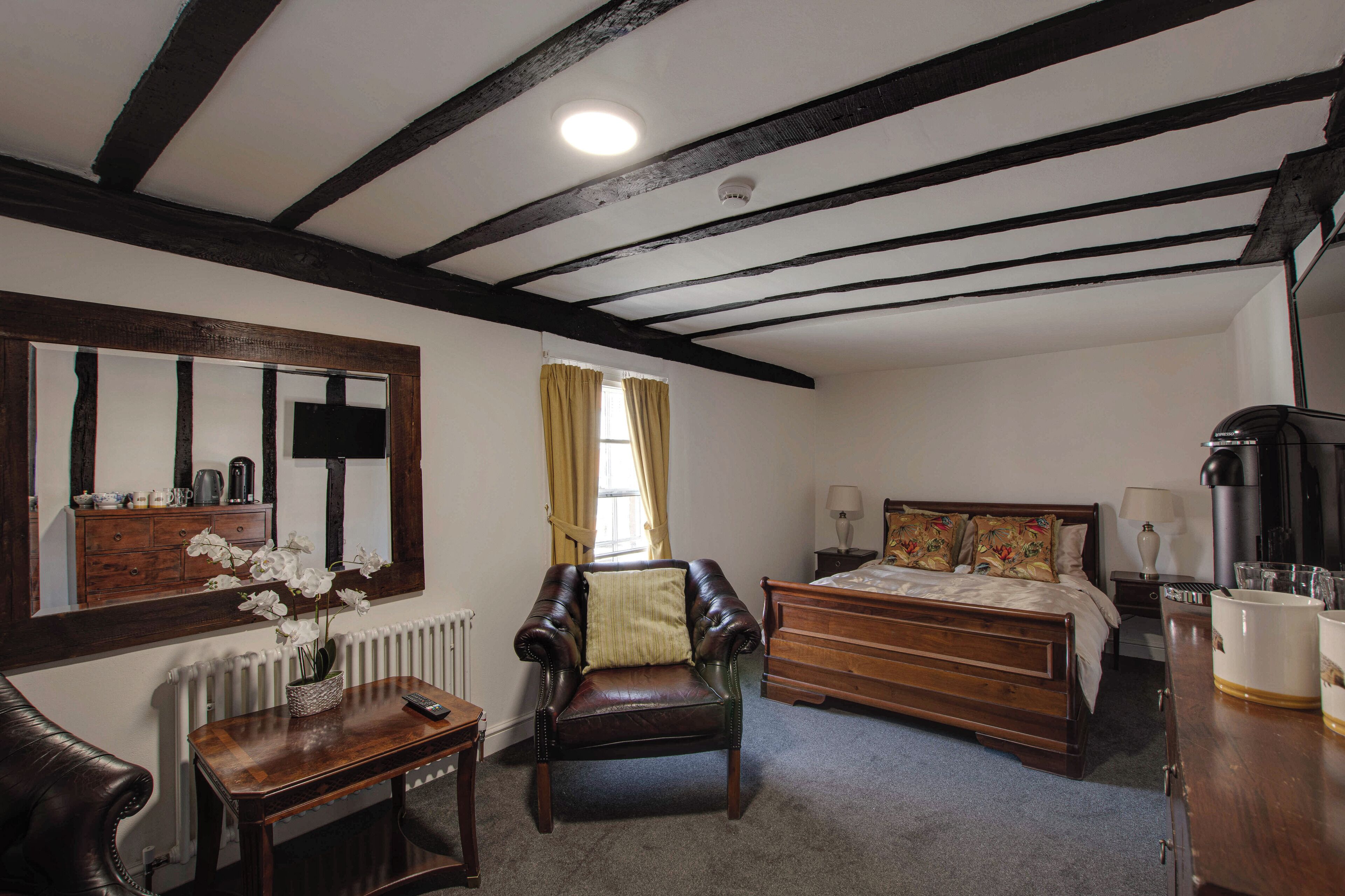 A room with a wooden bed, a chair, a table with a vase of flowers, a mirror, and a radiator.
