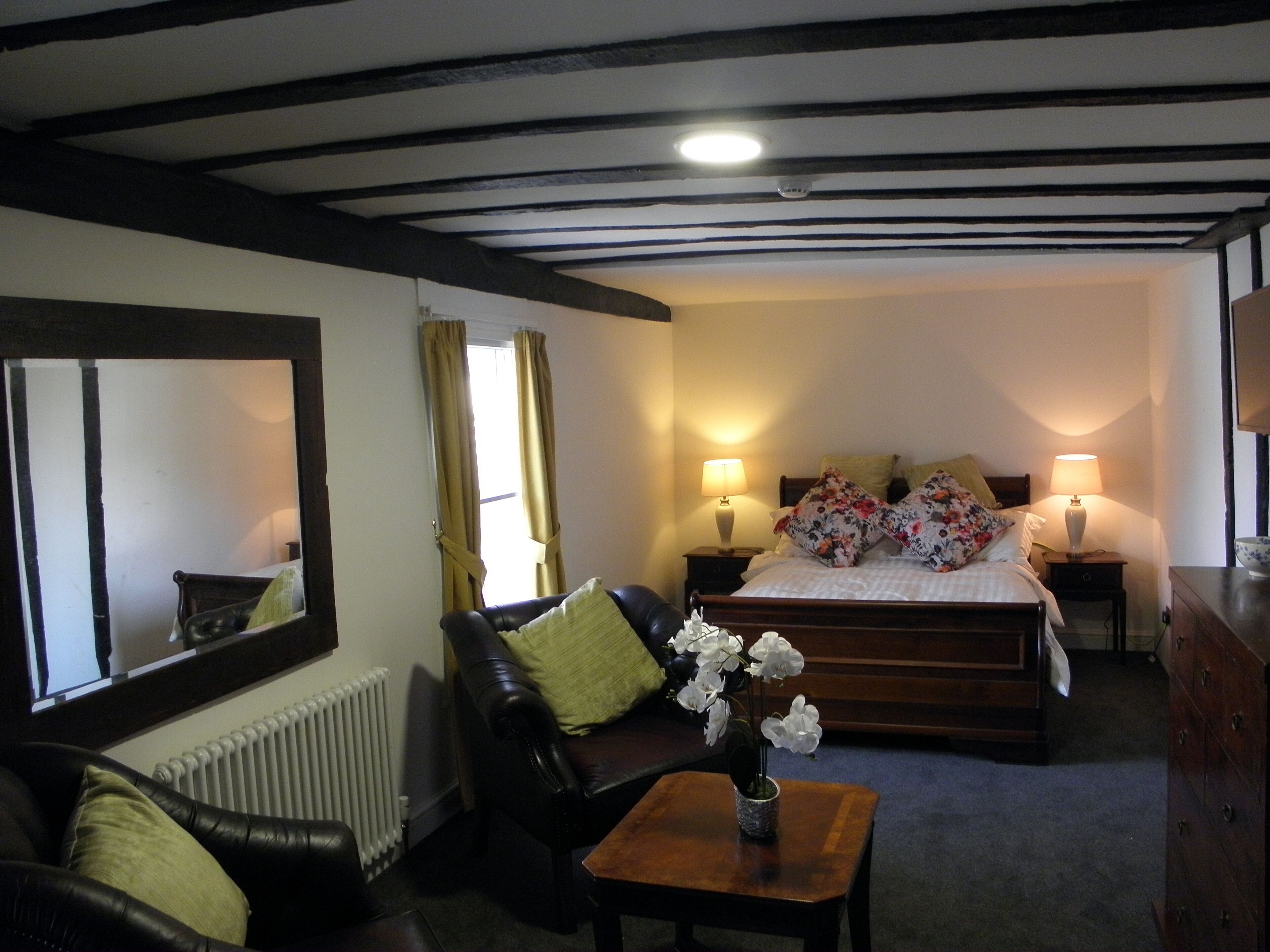 A room with a bed, two bedside lamps, a mirror, a radiator, a leather sofa, and a small table with a vase of flowers.