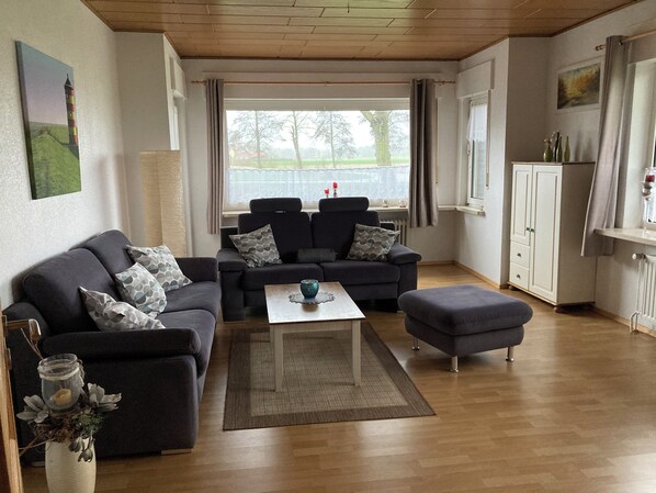 A living room with a sofa set, a coffee table, and a view of a lake through large windows. - Spacious vacation home that can accommodate up to 8 people, located very close to the North Sea coast (Großheide)