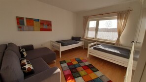 A small, well-lit room with a gray sofa, two single beds, a colorful rug, and a window with curtains. - Spacious vacation home that can accommodate up to 8 people, located very close to the North Sea coast (Großheide)