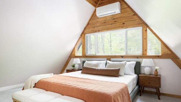 Un dormitorio con techo inclinado, un ventanal grande, una cama con muchos cojines, mesitas de noche con lámparas y un banco.