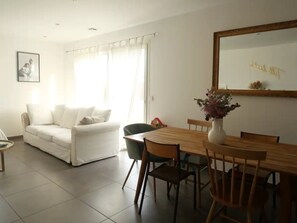 Una sala de estar con un sofá blanco, una mesa de comedor con sillas, un jarrón con flores y un espejo grande en la pared.