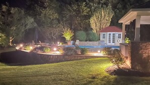 Et veloplyst udendørsområde med en mindre bygning, muligvis en pavillon, omgivet af en anlagt have med forskellige planter og græsplæne.