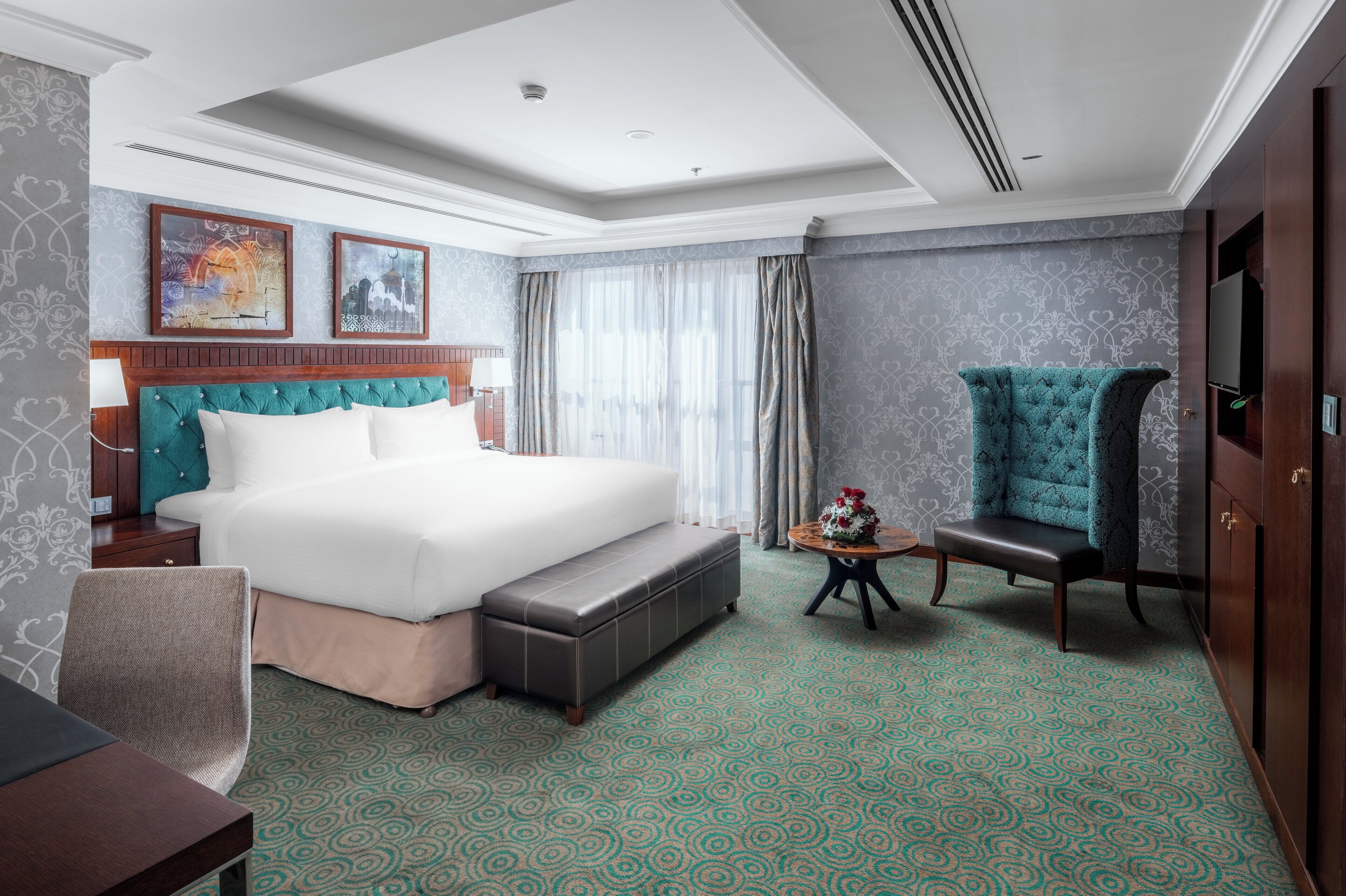 A hotel room with a large bed, a bench, a chair, a small table with a vase, and a TV.