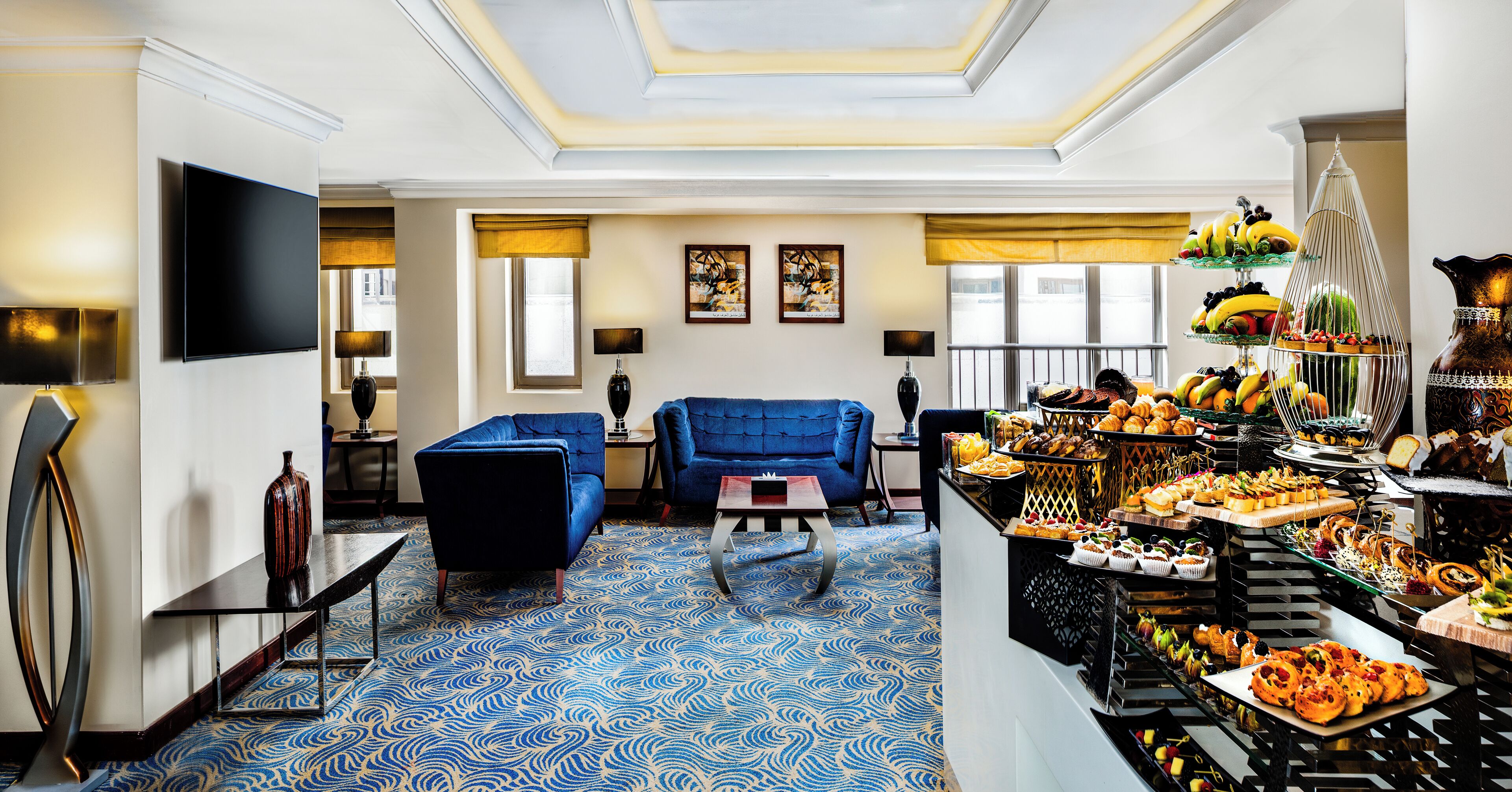 Um hall de hotel com carpete azul estampado, uma área de estar com sofás azuis, um buffet com diversos alimentos e obras de arte nas paredes.