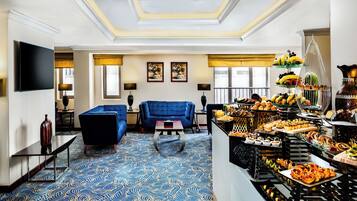 Um hall de hotel com carpete azul estampado, uma área de estar com sofás azuis, um buffet com diversos alimentos e obras de arte nas paredes.