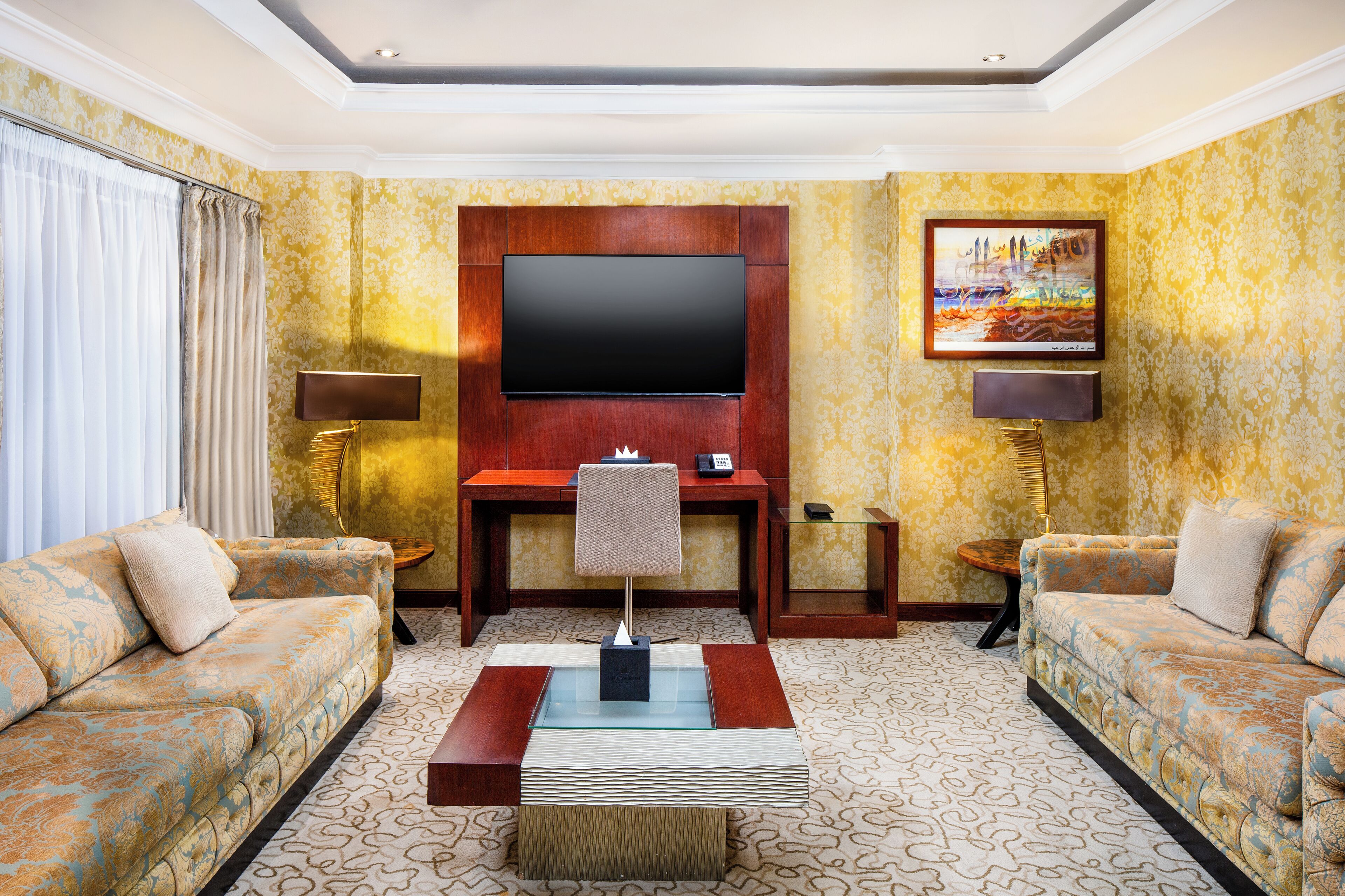 Habitación de hotel con un televisor de pantalla plana grande montado sobre un panel de madera, dos sofás, una mesa de centro y una alfombra estampada.