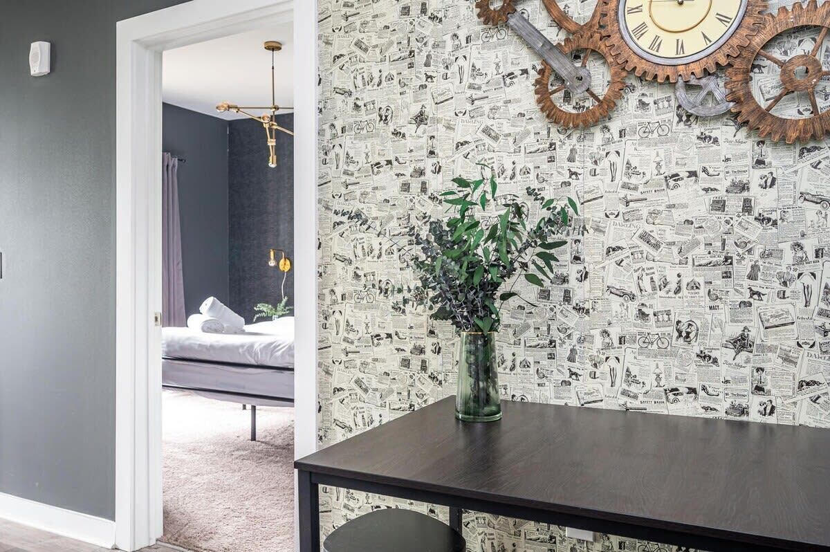 Een kamer met een donkergrijze muur, een houten klok en behangpapier met een krantenprint.