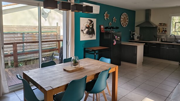 Una cocina moderna con comedor, que incluye una mesa de madera y sillas color turquesa, un refrigerador negro y un amplio ventanal con vista a una terraza de madera.