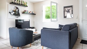 Una sala de estar moderna con un sofá y un sillón grises, un televisor de pantalla plana, una mesa de centro y una estantería con botellas de vino.