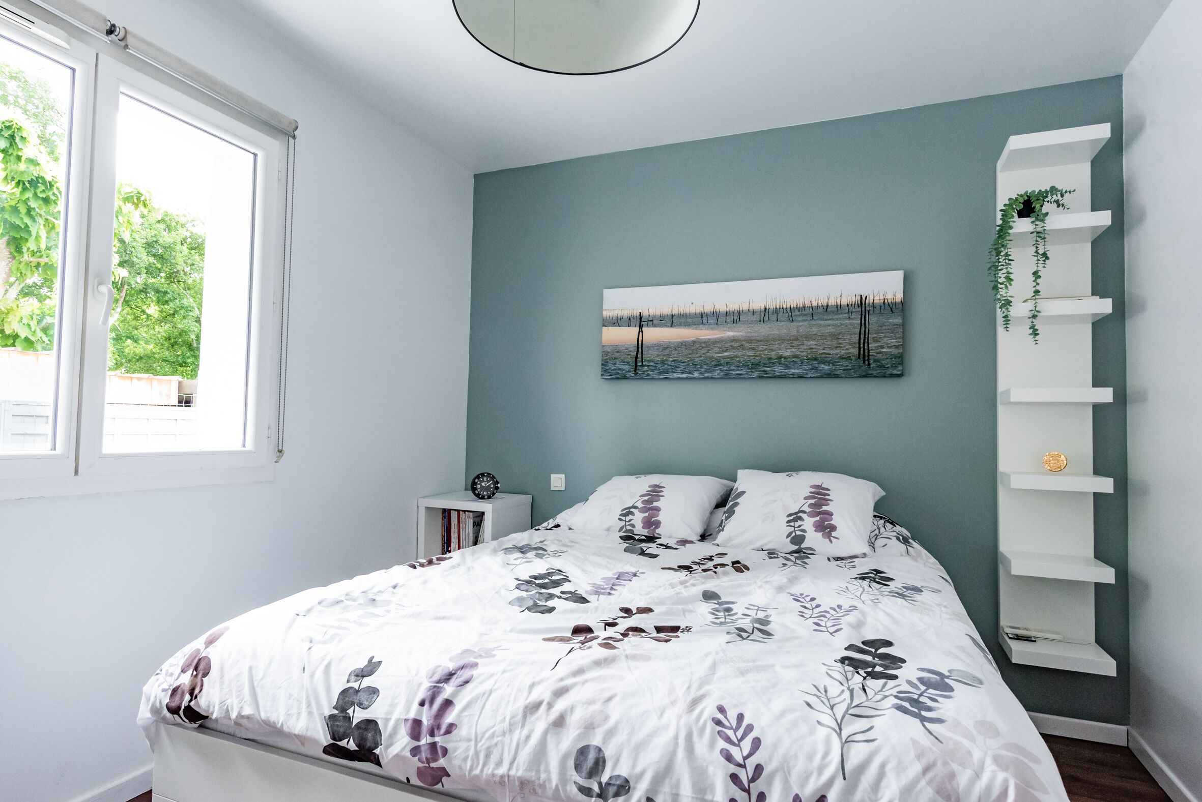 A bedroom with a bed, a nightstand, a wall-mounted shelf, and a window.