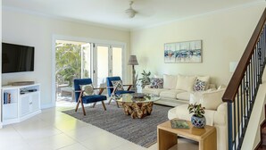A living room with a TV, blue chairs, a white sofa, a glass coffee table, and a staircase.