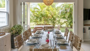 A dining area with a table set for six, surrounded by wicker chairs, a large potted plant, and a view of palm trees through open doors.