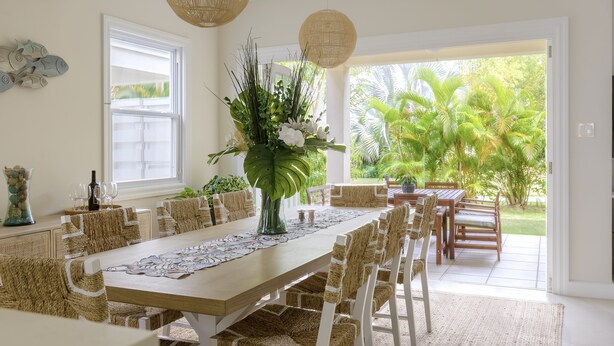 A spacious dining area with a long wooden table, woven chairs, and a large vase of flowers.