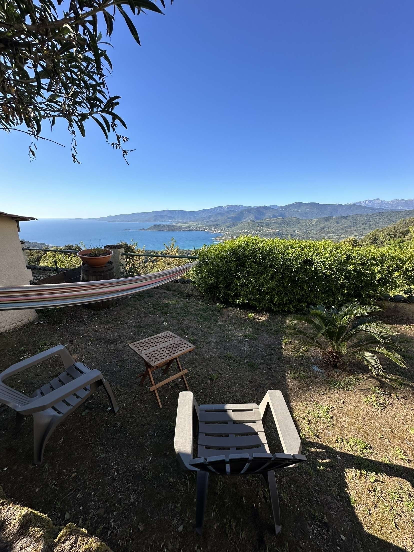 A patio with two chairs, a small table, and a hammock overlooking a scenic view of mountains and the sea.