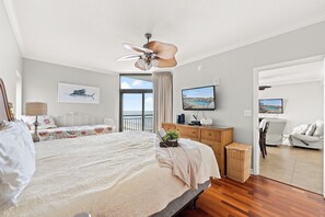 A bedroom with a bed, a ceiling fan, a television, and a view of the sea through a window.